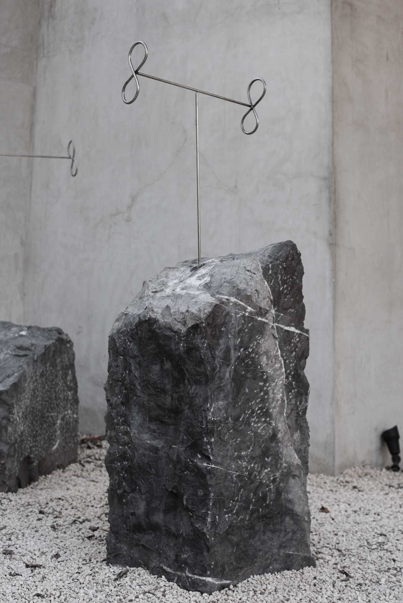 Sculpture II: an upright dark marble block on white gravel, steel post with two figure-eight loops, against concrete
