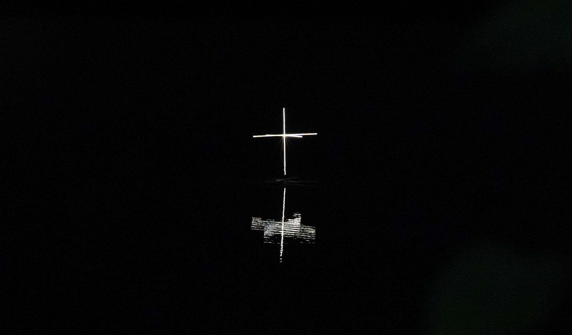 A single cross of light on black water, reflection below